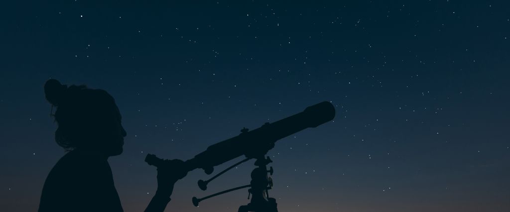 Silhouette of a person looking through a telescope against a starry night sky